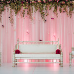 This Pakistani wedding showcases a white stage/flooring, blush draped backdrop with glass beaded curtains and hanging teardrop vases, and a cascading fresh floral band.