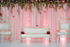 This Pakistani wedding showcases a white stage/flooring, blush draped backdrop with glass beaded curtains and hanging teardrop vases, and a cascading fresh floral band.